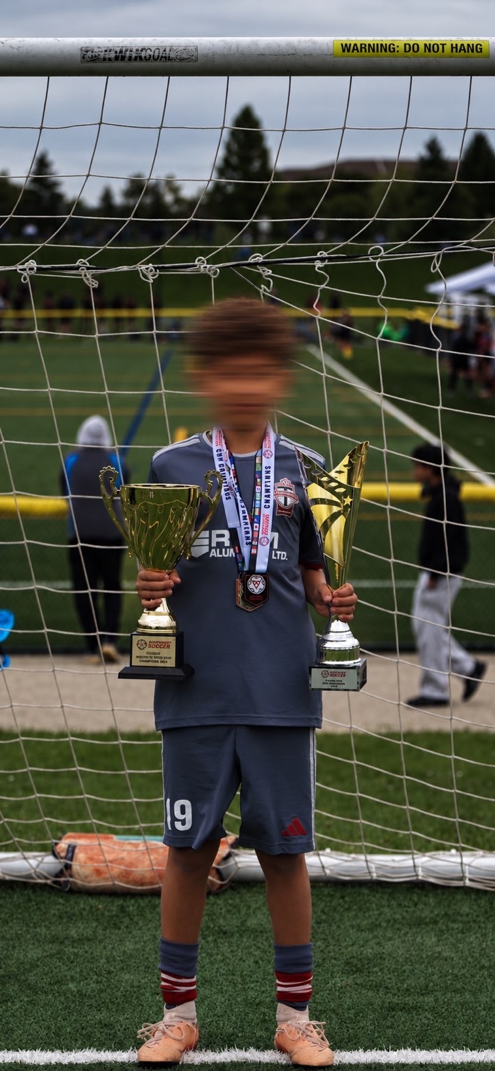 Zain Al Haj Ali standing in the goal mouth holding two championship trophies after winning the title.