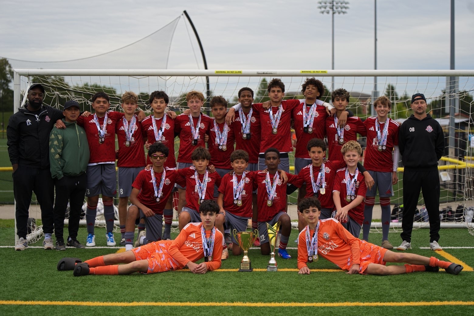 The team in red championship kits posing with the trophy and coaching staff.